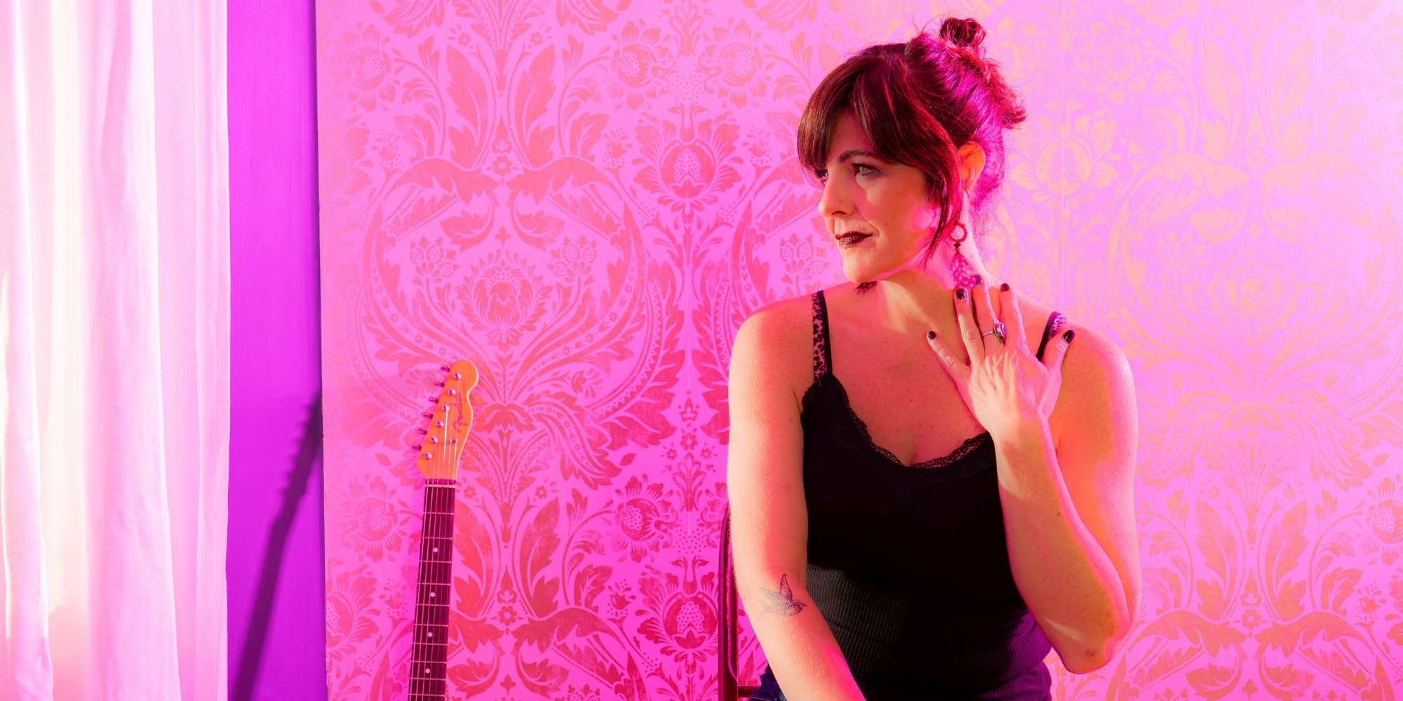 Woman seated beside a guitar in a pink-lit room with patterned wallpaper, looking to the side.